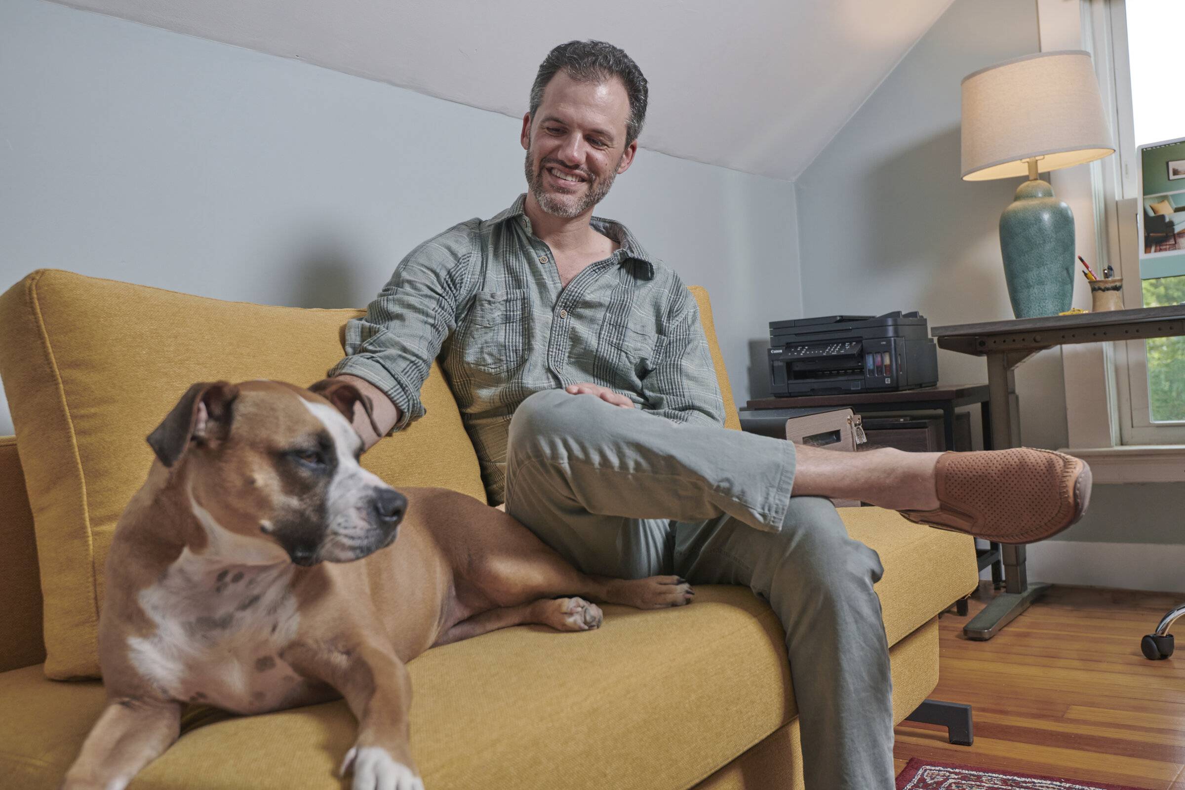 Marcus Berley on a yellow couch at home, smiling, with his dog beside him